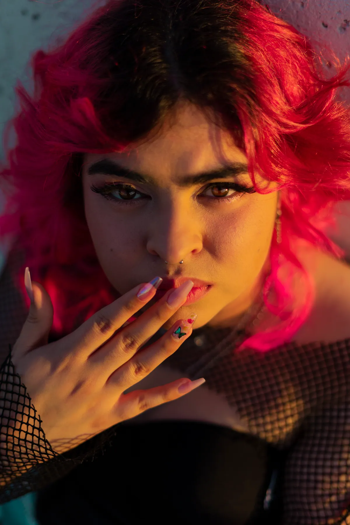 Close-up portrait of a person with vivid pink hair, hand raised to their lips