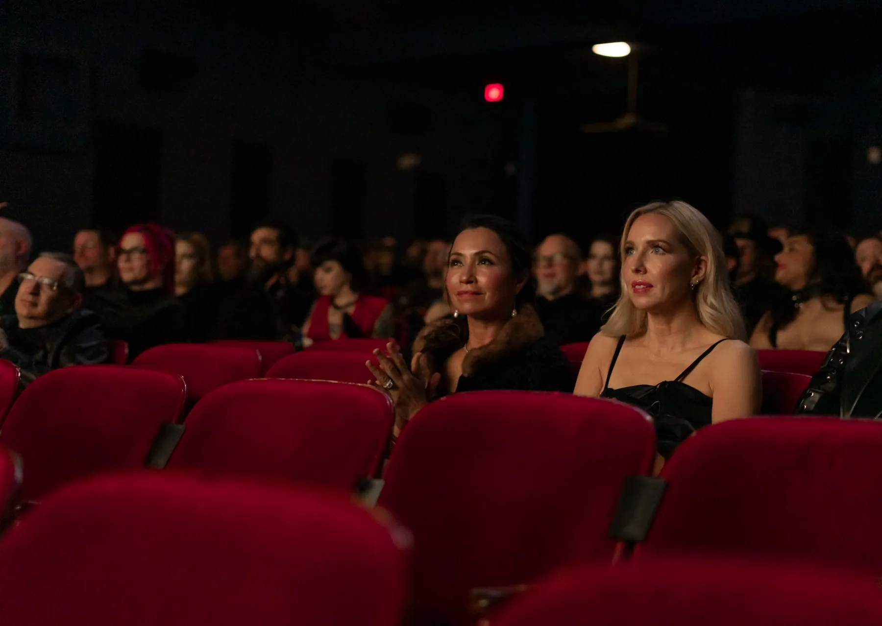 Audience seated in red velvet theatre seats, faces softly lit in the dark