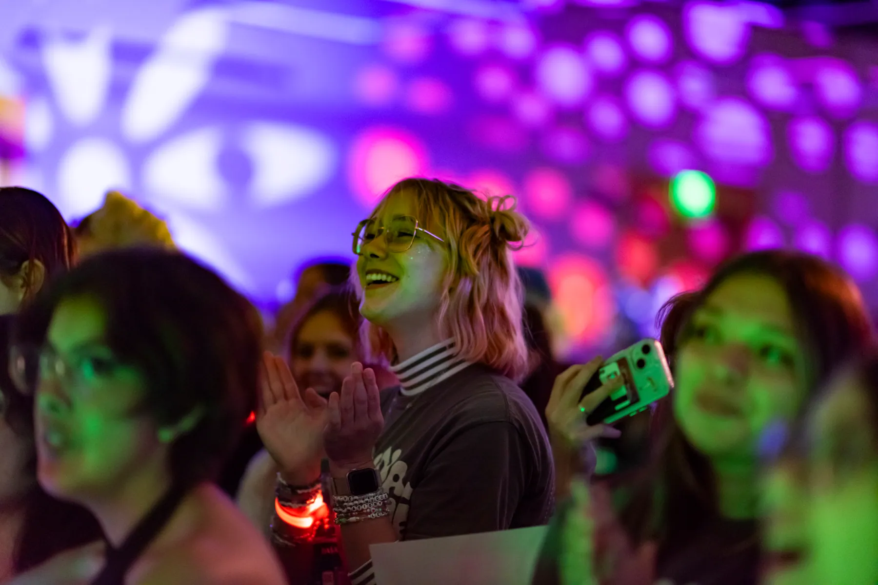 Concert crowd in purple and green light, one person in glasses smiling and clapping