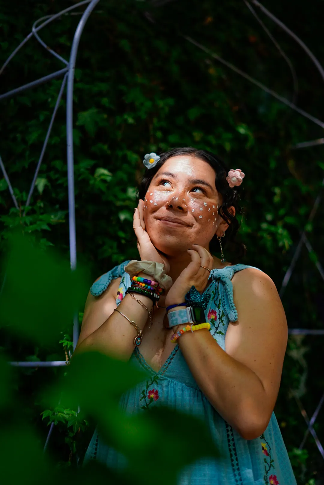 Person in a blue embroidered dress with flowers in her hair, hands on her cheeks, looking upward