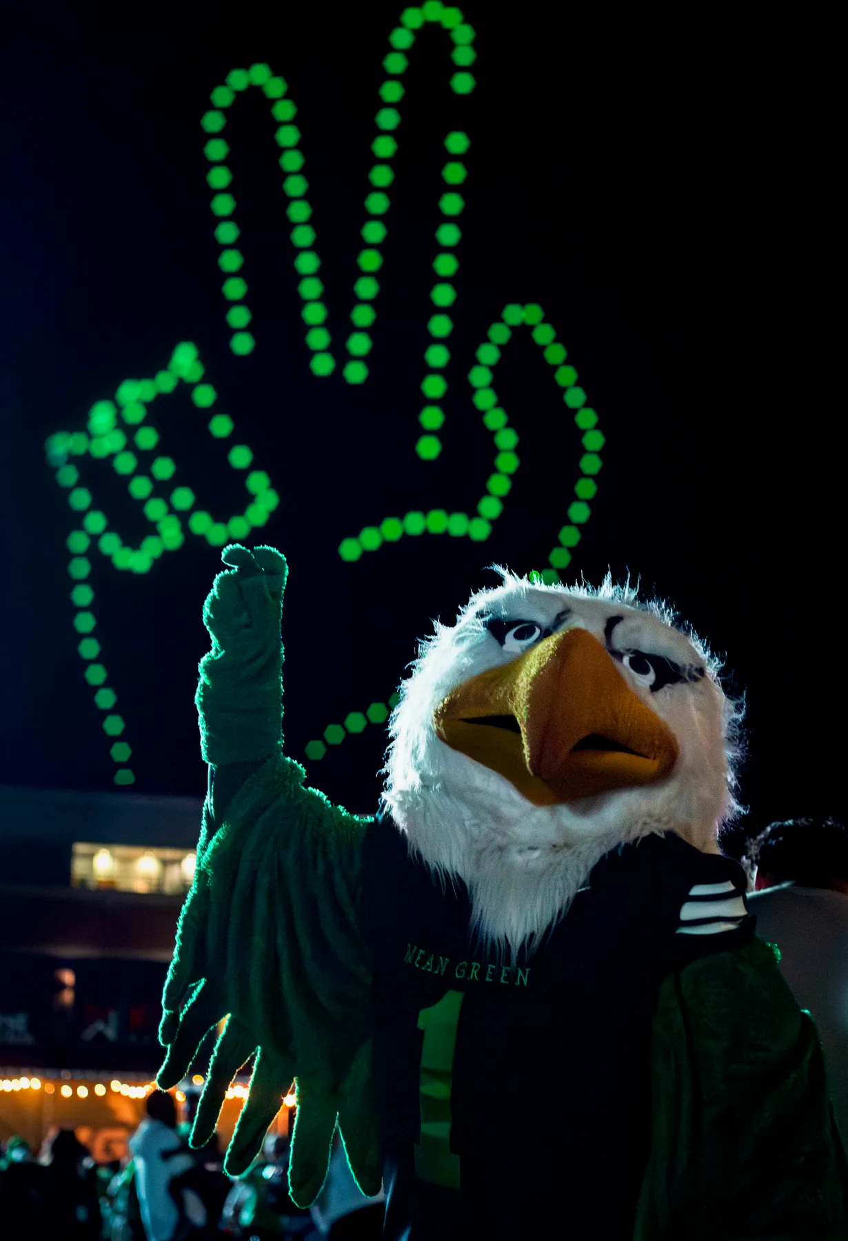 Eagle mascot in a green jersey waving beneath a glowing drone-light hand sign at night