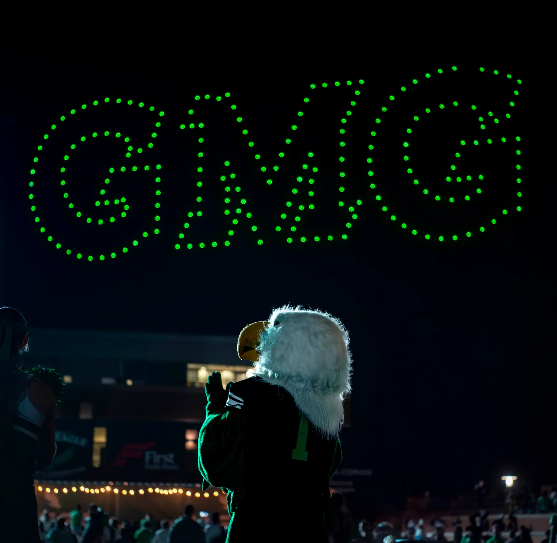 Eagle mascot seen from behind beneath green drone lights spelling GMG at night