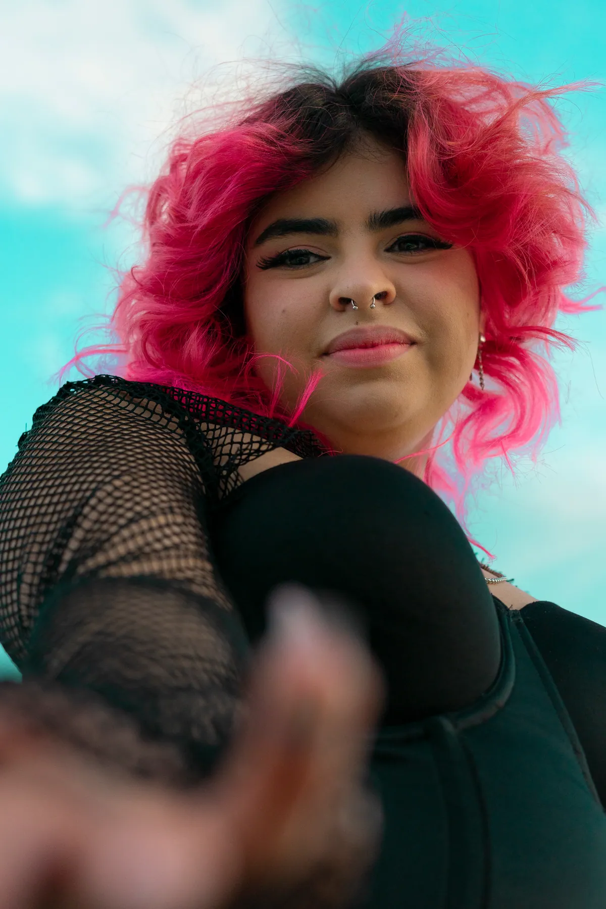 Low-angle portrait of a person with pink hair reaching toward the camera against a bright blue sky